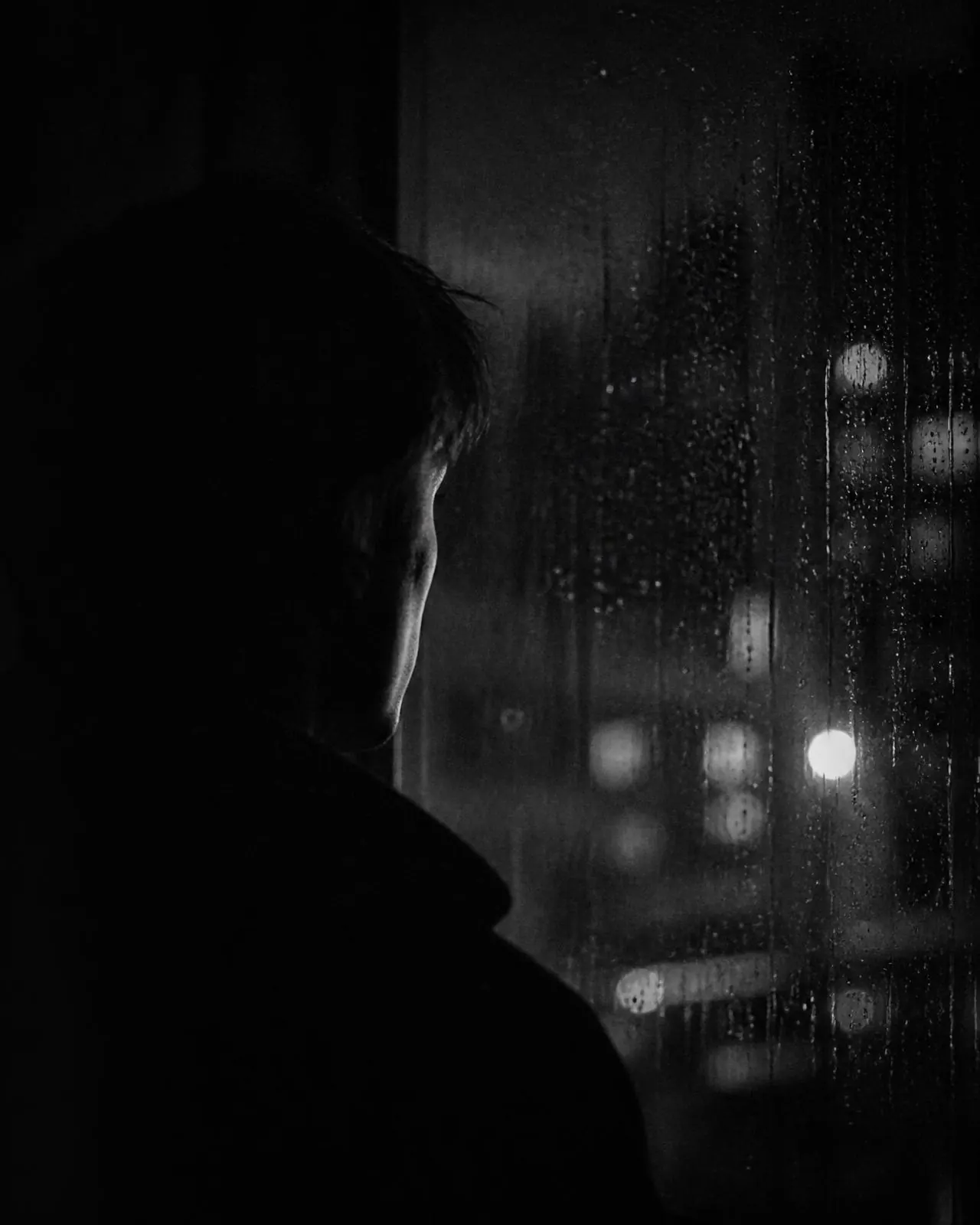 A figure in shadow watching city lights through a rain covered window at night